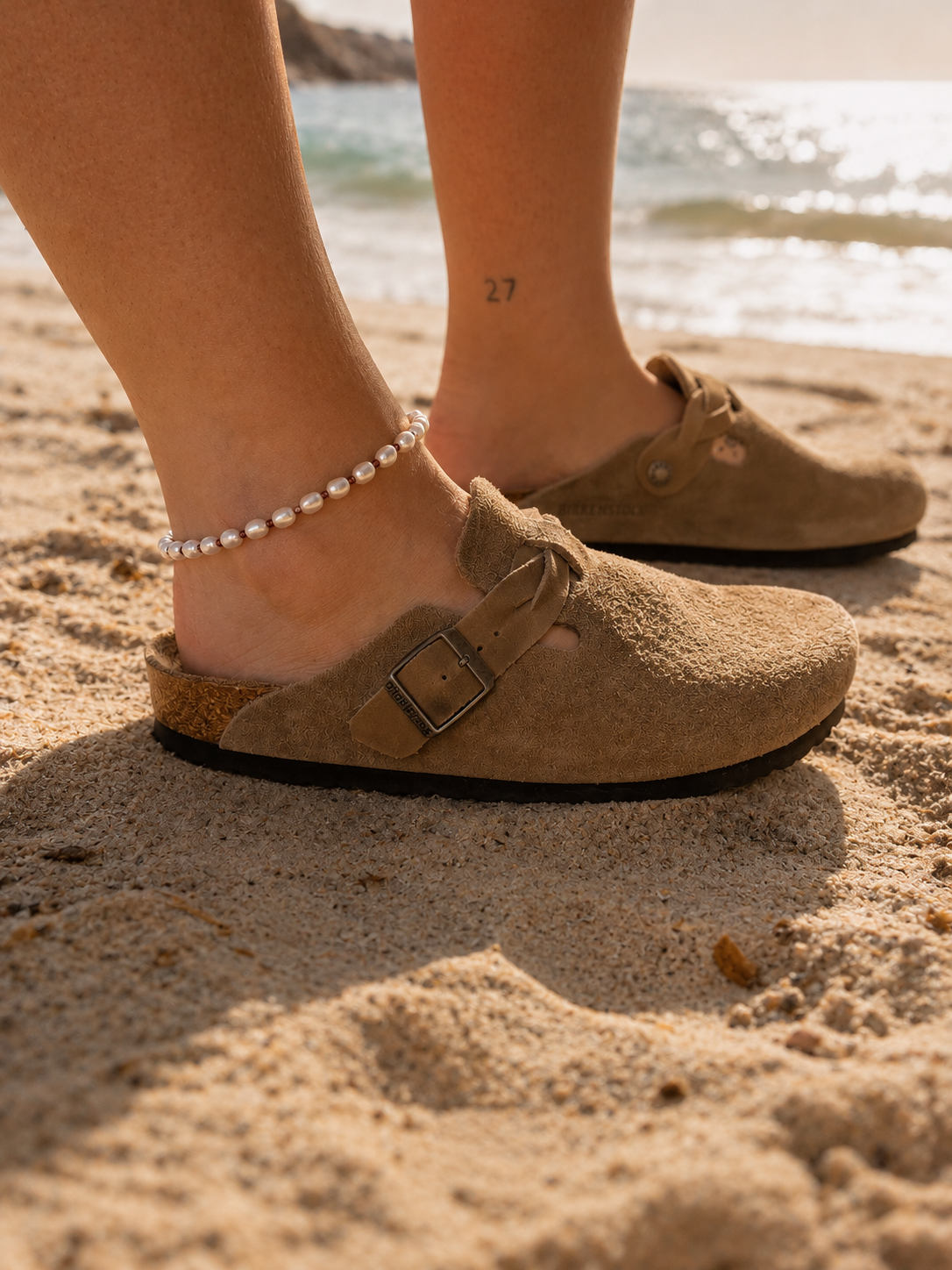 ChatGPT Image 17 mai 2026 22_31_51 | MAISON LIAMA Close-up of tan cork sandals on sandy beach, with a pearl ankle bracelet and a small '27' tattoo on the ankle, ocean in the background.