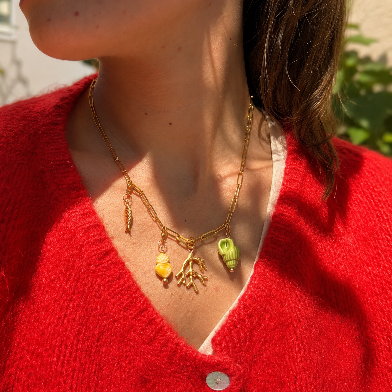 ChatGPT Image 17 mai 2026 23_57_02 | MAISON LIAMA Gold chain necklace with sea-themed charms (fish, coral, shells) resting on a red knit cardigan.