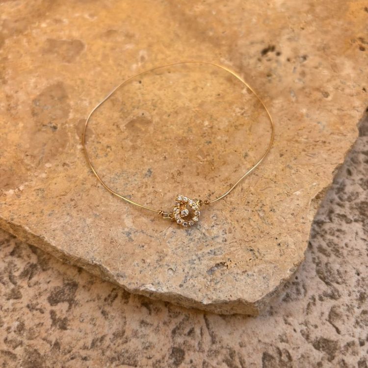 Gold bangle bracelet with a circular diamond-encrusted charm resting on a rough beige rock surface.