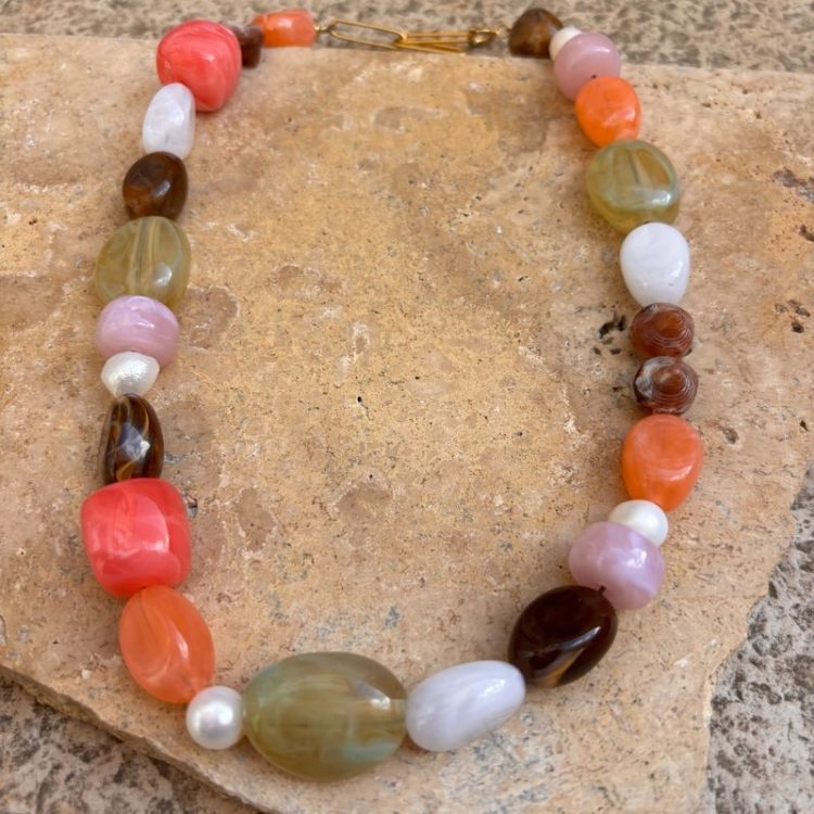 Colorful beaded necklace laid on a stone surface with large oval beads in coral, pink, lavender, white, green, and amber tones.