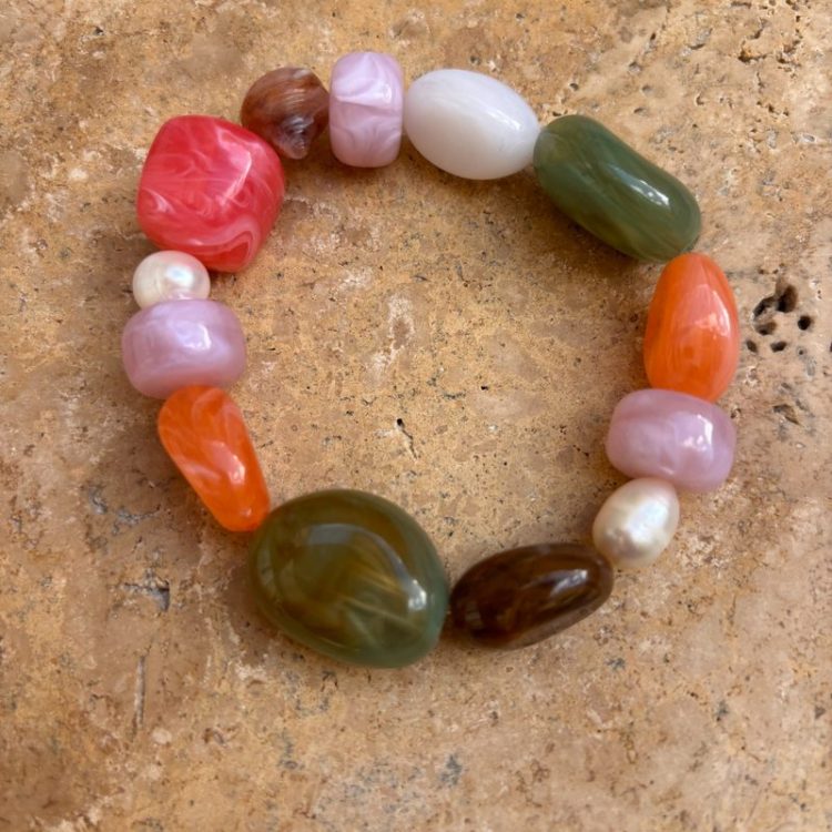 Colorful beaded bracelet arranged in a circle on a textured stone surface, featuring pink, red, orange, green, purple, and pearl beads.