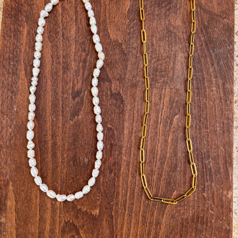 IMG_8361_800x800 | MAISON LIAMA Two necklaces lie on a wooden surface: a strand of irregular white pearls on the left and a long gold chain on the right.