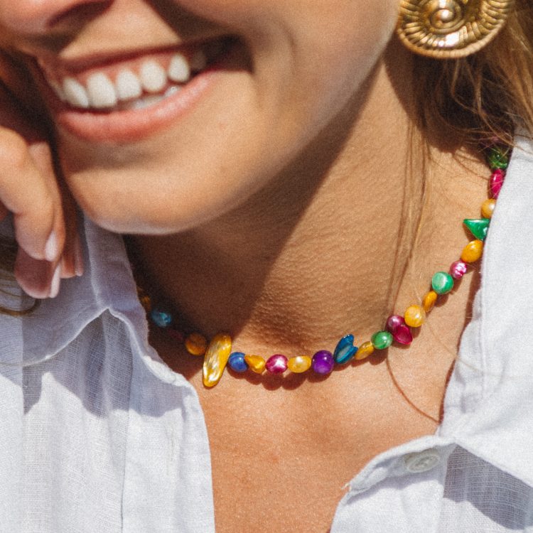 Close-up of a smiling woman’s face showing a gold spiral earring and colorful beaded necklace with a white shirt.