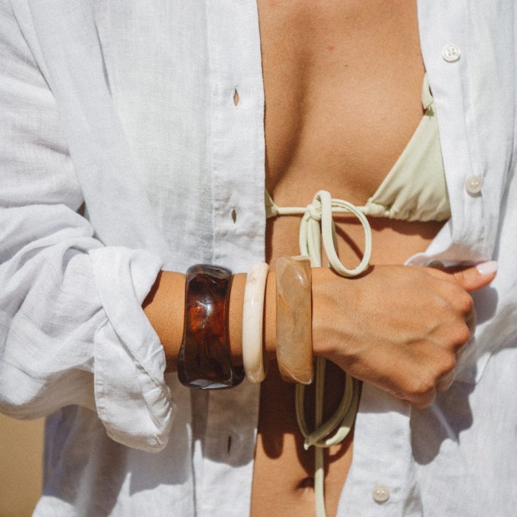 Person wearing an open white shirt over a light green bikini top, accessorized with chunky stone bracelets on the forearm.